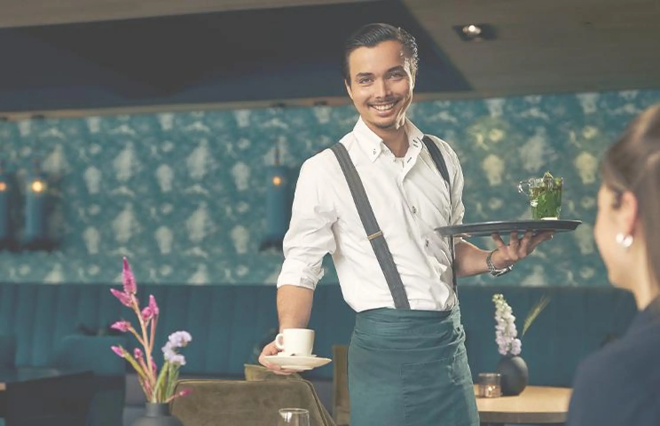 Medewerker bediening serveert kop koffie aan gast en kijkt lachend in camera